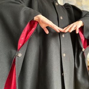 Pode incluir: Um casaco preto de mistura de lã com gola alta, fecho frontal com botões metálicos e mangas largas. O forro interior é de tecido vermelho vibrante decorado com um pequeno padrão repetitivo de folhas verde-escuras.