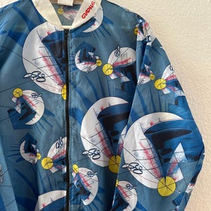 May include: A blue and white zip-up jacket with a geometric pattern. The jacket features a white collar and the word "GRAPHIA" in red on the collar. The pattern includes white, red, and yellow shapes on a blue background.