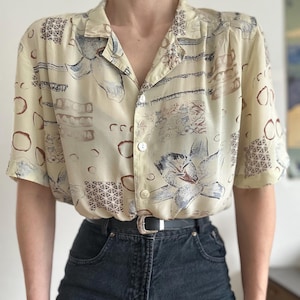 Puede incluir: Una camisa abotonada de color crema con un estampado floral en tonos azules, marrones y beige. La camisa tiene mangas cortas y se combina con vaqueros oscuros y un cinturón negro con hebilla plateada.