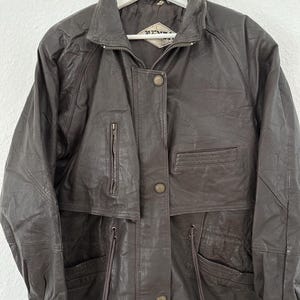 Puede incluir: Chaqueta de cuero marr&oacute;n oscuro con cuello, cremallera y botones. La chaqueta tiene m&uacute;ltiples bolsillos y una cintura con cord&oacute;n. Cuelga de una percha blanca.