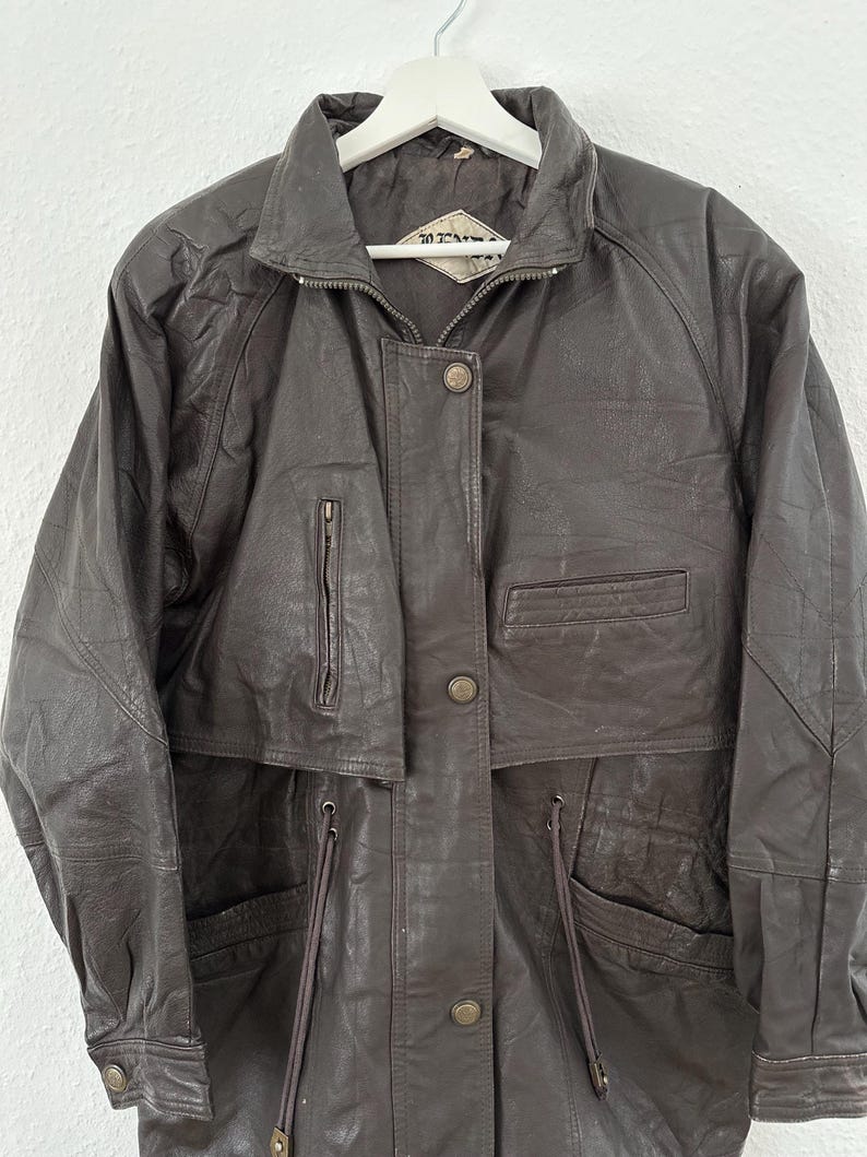 Puede incluir: Chaqueta de cuero marr&oacute;n oscuro con cuello, cremallera y botones. La chaqueta tiene m&uacute;ltiples bolsillos y una cintura con cord&oacute;n. Cuelga de una percha blanca.