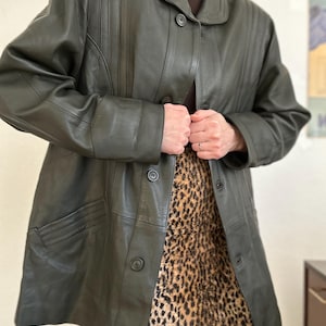 May include: Dark green leather coat with a button-up front, a collar, and vertical stitched detailing on the shoulders. The coat is worn over a leopard print skirt.