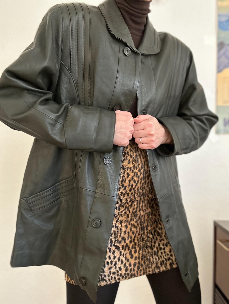 May include: Dark green leather coat with a button-up front, a collar, and vertical stitched detailing on the shoulders. The coat is worn over a leopard print skirt.