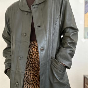 May include: Olive green leather coat with a button-up front, a rounded collar, and vertical stitched details. The coat has two pockets and is worn over a brown turtleneck and a leopard print skirt.
