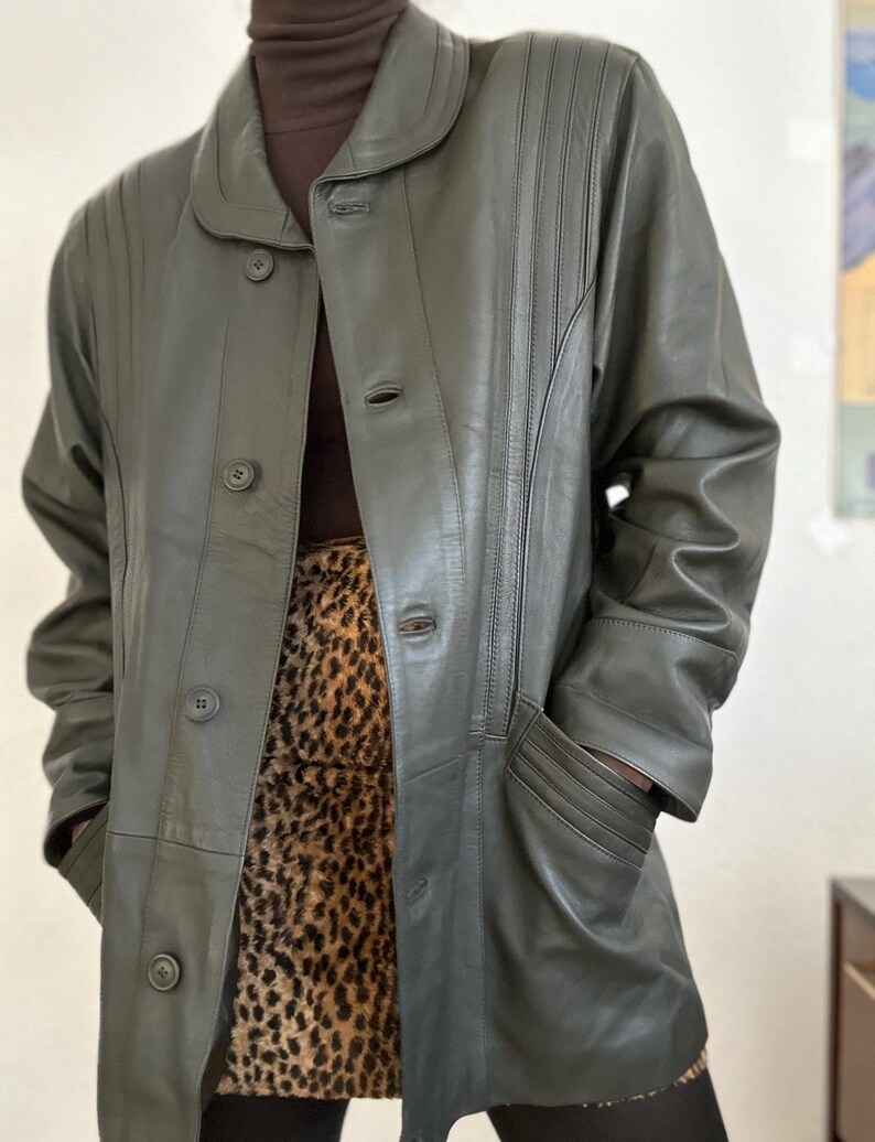 May include: Olive green leather coat with a button-up front, a rounded collar, and vertical stitched details. The coat has two pockets and is worn over a brown turtleneck and a leopard print skirt.