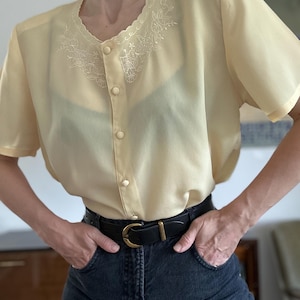 May include: A pale yellow short-sleeved blouse featuring a scalloped neckline with white floral embroidery, a front button placket with matching yellow buttons, and a semi-sheer fabric texture, paired with dark charcoal denim jeans and a black belt.