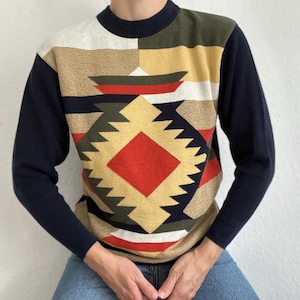 May include: A long-sleeved sweater with a geometric pattern in shades of gold, red, green, and navy blue. The sweater has a navy blue collar and sleeves. The design features a central diamond shape and zigzag edges.
