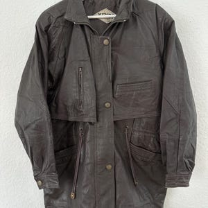 Puede incluir: Chaqueta de cuero marr&oacute;n oscuro con cremallera, botones y m&uacute;ltiples bolsillos. La chaqueta tiene cuello y mangas largas. Cuelga de una percha blanca.