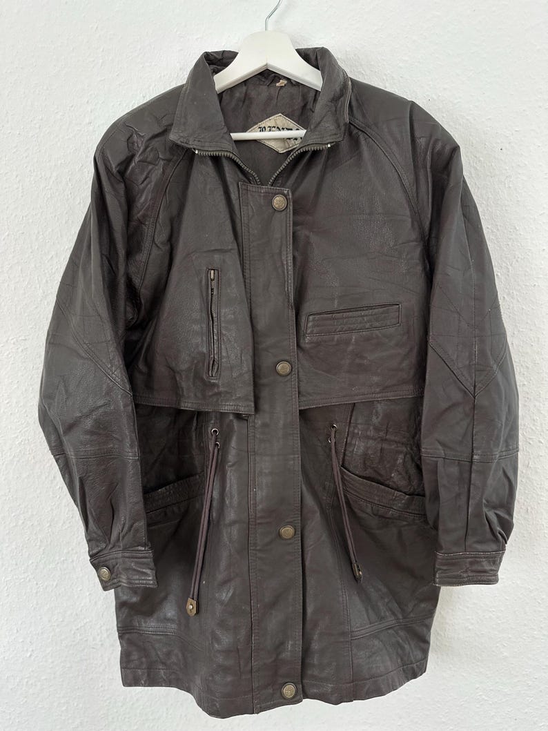 Puede incluir: Chaqueta de cuero marr&oacute;n oscuro con cremallera, botones y m&uacute;ltiples bolsillos. La chaqueta tiene cuello y mangas largas. Cuelga de una percha blanca.