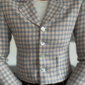 May include: A cropped blazer featuring a multi-colored plaid pattern in shades of blue, peach, and cream. The garment has a notched lapel, long sleeves, and a front closure with four iridescent, round buttons. Two small pockets are visible at the waist.
