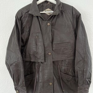 Puede incluir: Chaqueta de cuero marr&oacute;n con cuello alzado, cremallera y botones. La chaqueta tiene m&uacute;ltiples bolsillos y cintura con cord&oacute;n. La chaqueta est&aacute; colgada de una percha blanca.
