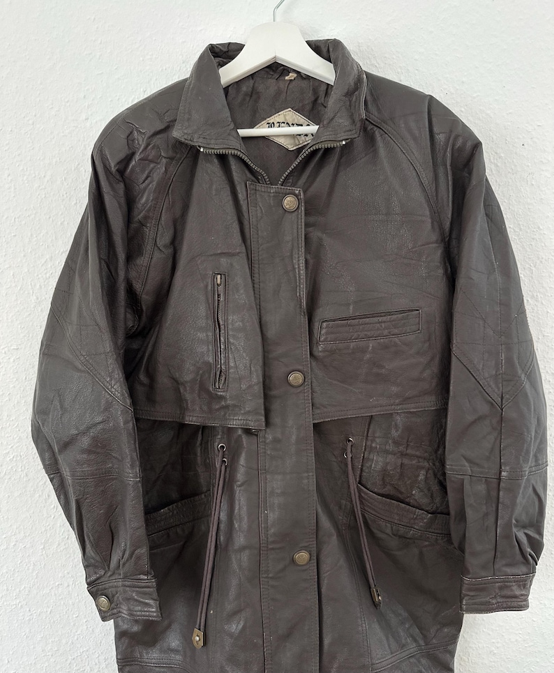 Puede incluir: Chaqueta de cuero marr&oacute;n con cuello alzado, cremallera y botones. La chaqueta tiene m&uacute;ltiples bolsillos y cintura con cord&oacute;n. La chaqueta est&aacute; colgada de una percha blanca.