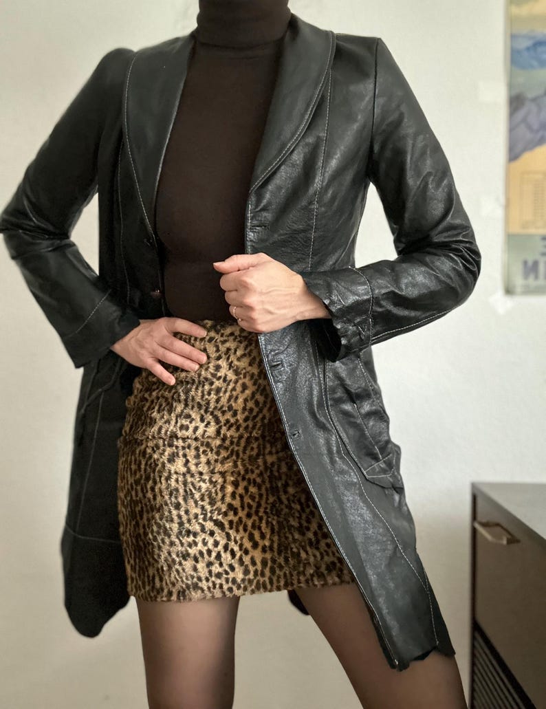 May include: A black leather jacket with a notched collar and a button closure. The jacket is worn over a brown turtleneck and a leopard print skirt. The outfit is completed with black tights.