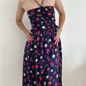 Puede incluir: Vestido azul marino con estampado floral con flores rojas, rosas y blancas. El vestido tiene un corpiño sin tirantes con tirantes finos y un detalle fruncido. La falda llega a la rodilla y también presenta el estampado floral.