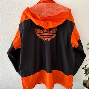 Può includere: Giacca Adidas vintage arancione e nera con cappuccio. La giacca presenta il logo Adidas arancione sul retro. La giacca è in stile antivento con chiusura a cerniera.