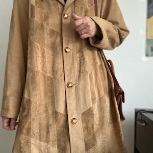 Puede incluir: Un abrigo de ante color beige con cuello y cierre de botones. El abrigo presenta un diseño de patchwork con patrones verticales y en zigzag. Se ve un bolso de hombro de cuero marrón.