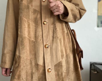 Könnte beinhalten: Ein beigefarbener Wildledermantel mit Kragen und Knopfverschluss. Der Mantel hat ein Patchwork-Design mit vertikalen und Zickzackmustern. Eine braune Leder-Umhängetasche ist sichtbar.