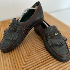 Puede incluir: Un par de mocasines de cuero en tonos grises y marrones. Los zapatos presentan un detalle de flecos y un emblema decorativo en la pala. Las suelas y los ribetes son de color marr&oacute;n oscuro. Se exhiben sobre una superficie de madera.