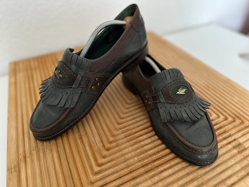 Puede incluir: Un par de mocasines de cuero en tonos grises y marrones. Los zapatos presentan un detalle de flecos y un emblema decorativo en la pala. Las suelas y los ribetes son de color marr&oacute;n oscuro. Se exhiben sobre una superficie de madera.