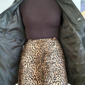 May include: An open, dark green leather jacket with two buttons, revealing a brown turtleneck and a leopard print skirt. The jacket's interior features two pockets. The skirt has a brown and black spotted pattern.