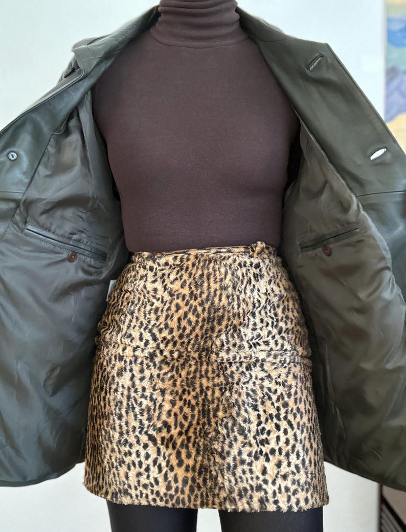 May include: An open, dark green leather jacket with two buttons, revealing a brown turtleneck and a leopard print skirt. The jacket's interior features two pockets. The skirt has a brown and black spotted pattern.