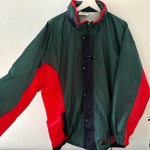 Könnte beinhalten: Eine grüne, rote und marineblaue Vintage-Jacke mit hohem Kragen und Reißverschluss. Die Jacke hat rote Einsätze an den Ärmeln und einen marineblauen Streifen auf der Vorderseite. Die Jacke hängt an einem weißen Kleiderbügel.