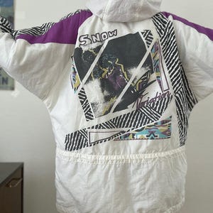 May include: White and purple vintage ski jacket with a hood. The back features a graphic design with the word "SNOW" and a geometric pattern. The jacket has black and white striped accents on the sleeves.