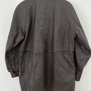 Puede incluir: Un abrigo de cuero marr&oacute;n oscuro con cierre de botones y cuello. El abrigo est&aacute; colgado de una percha de metal sobre un fondo blanco. El abrigo tiene una longitud larga y un dise&ntilde;o cl&aacute;sico.