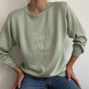 Puede incluir: Un suéter verde claro con cuello redondo y mangas largas. El suéter presenta un diseño floral en relieve en la parte delantera. La tela tiene un brillo sutil. La persona lleva vaqueros azules.