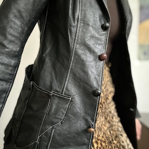 May include: Black leather jacket with white stitching details, featuring a notched collar, two front pockets, and three buttons. The jacket is paired with a leopard print skirt. The jacket's design includes a classic tailored silhouette.