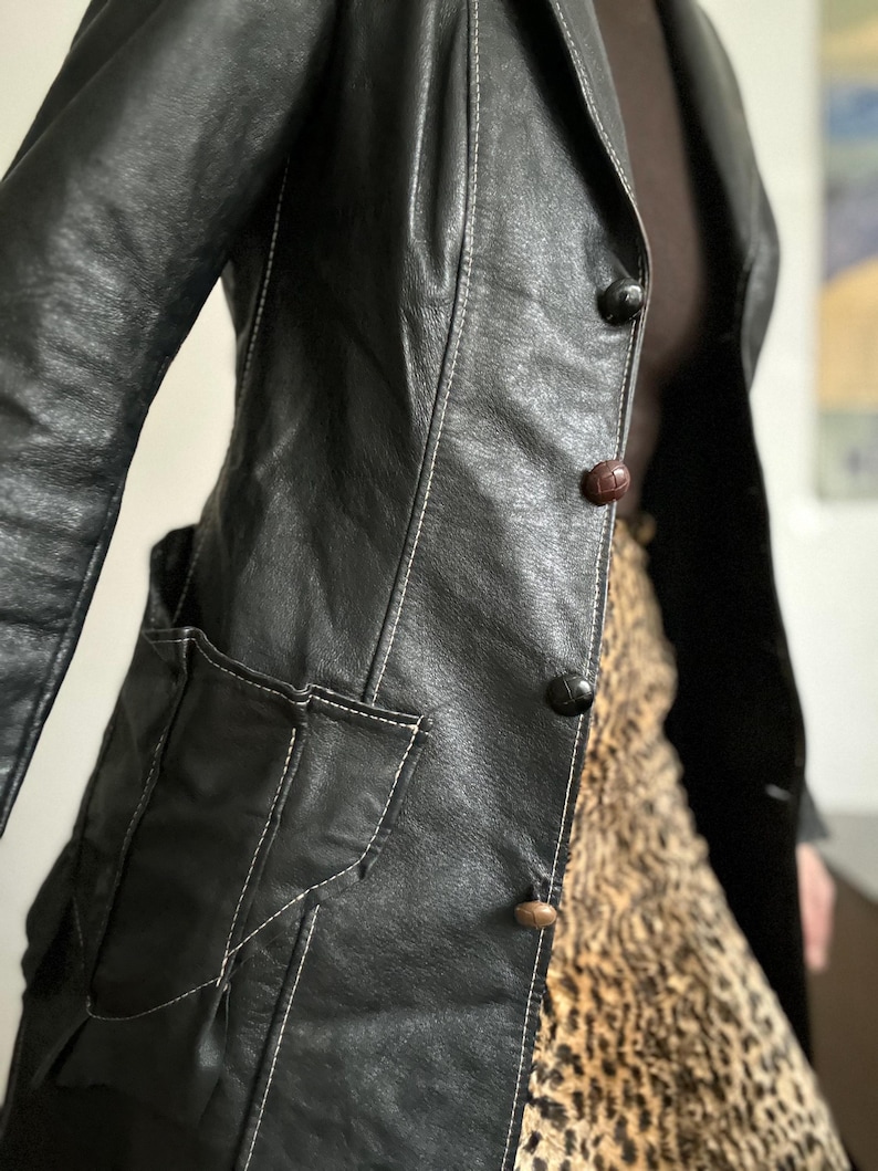May include: Black leather jacket with white stitching details, featuring a notched collar, two front pockets, and three buttons. The jacket is paired with a leopard print skirt. The jacket's design includes a classic tailored silhouette.