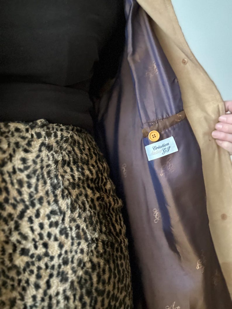 Może przedstawiać: Beżowy zamszowy płaszcz z ciemnofioletową podszewką. Płaszcz ma zapięcie na guziki i metkę z napisem "Creation Atelier Gf". Podszewka ma subtelny wz&oacute;r. Płaszcz jest otwarty, odsłaniając wnętrze.