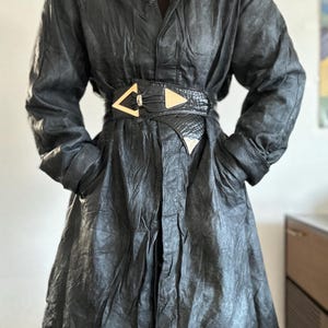 Peut inclure: Un manteau en cuir noir avec un col et des manches longues. Le manteau est cintré à la taille par une ceinture noire avec des accents géométriques dorés. Le manteau a une fermeture boutonnée et des poches.
