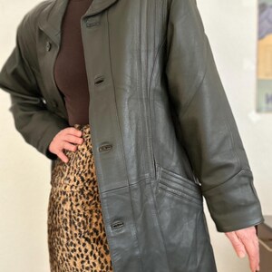 May include: Dark green leather jacket with a button-up front, a notched collar, and vertical stitched detailing on the front. The jacket has a long length and a tailored fit. The model is wearing a brown turtleneck and a leopard print skirt.