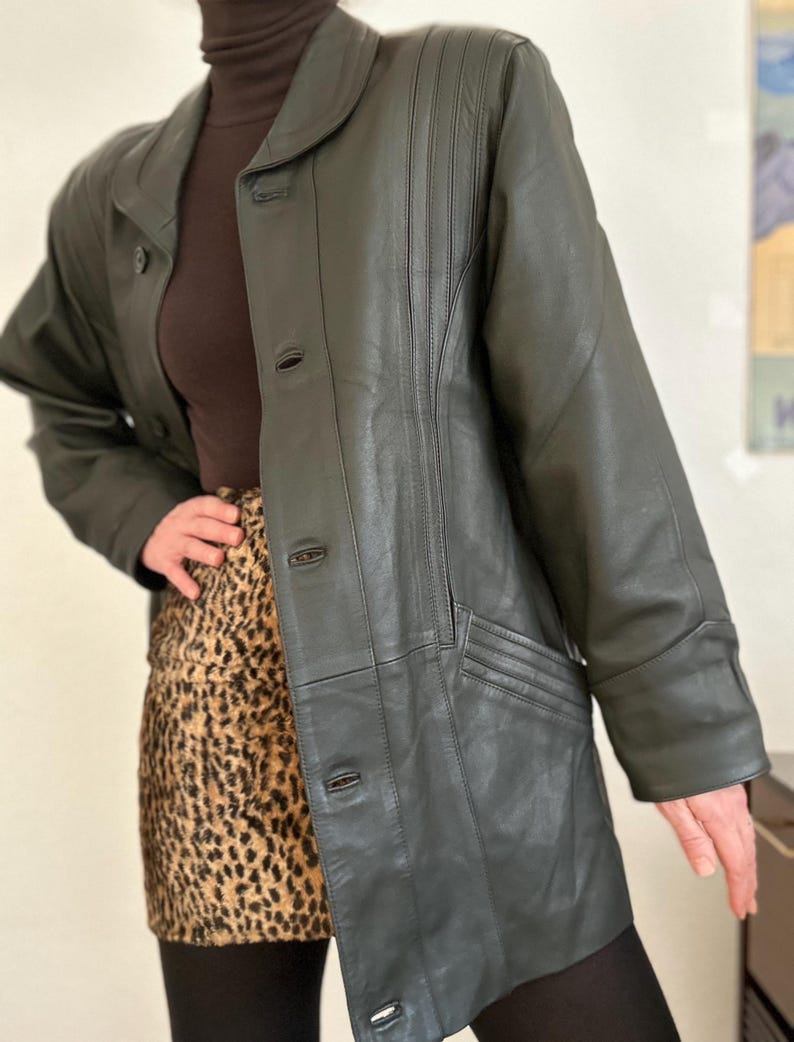 May include: Dark green leather jacket with a button-up front, a notched collar, and vertical stitched detailing on the front. The jacket has a long length and a tailored fit. The model is wearing a brown turtleneck and a leopard print skirt.