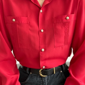 May include: A long-sleeved red button-down shirt featuring a pointed collar with vertical pleats, two chest pockets with vertical pleats and gold-toned buttons, and a central placket with matching gold-toned buttons, tucked into dark denim jeans.