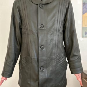 May include: Dark olive green leather jacket with a button-up front, a notched collar, and two front pockets. The jacket has vertical stitched details on the front and sleeves. The jacket is worn over a brown turtleneck.