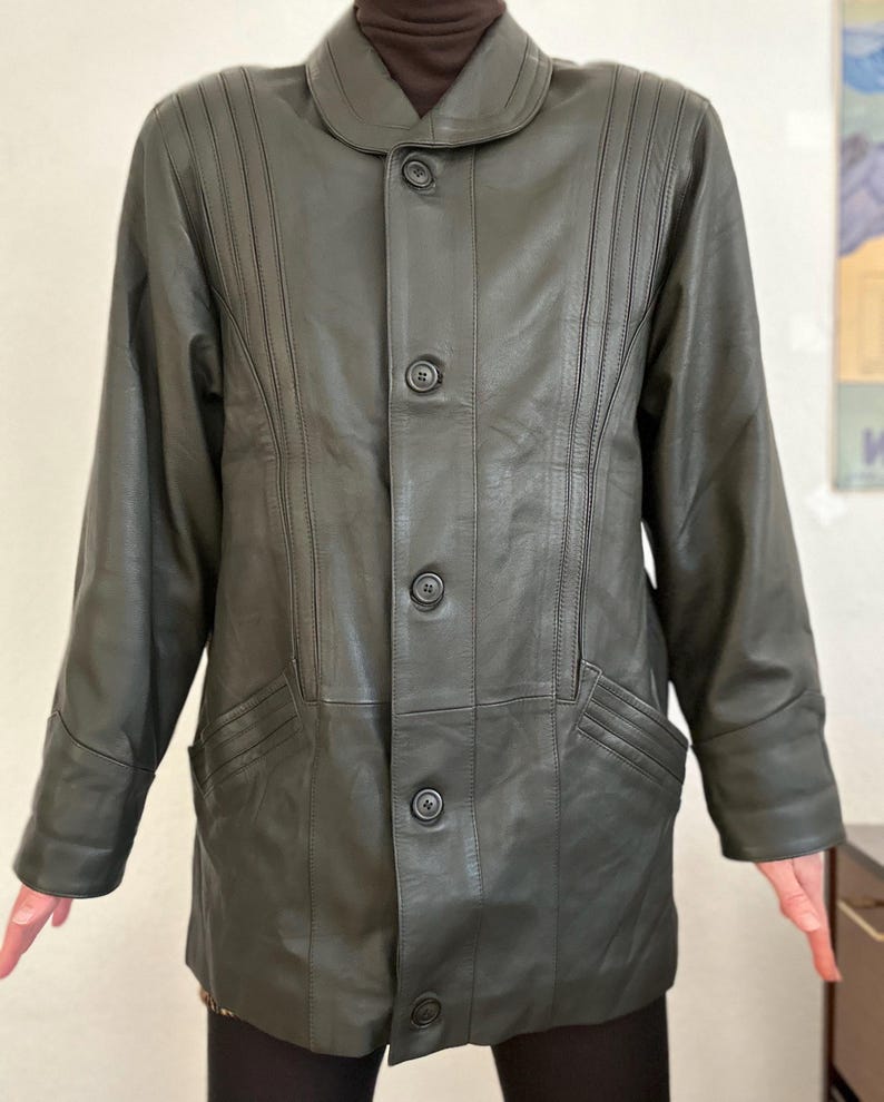 May include: Dark olive green leather jacket with a button-up front, a notched collar, and two front pockets. The jacket has vertical stitched details on the front and sleeves. The jacket is worn over a brown turtleneck.