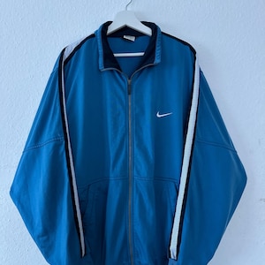 May include: A teal blue zip-up jacket with a white Nike logo on the chest. The jacket has black and white striped accents on the sleeves and a black collar. The jacket is hanging on a white hanger.