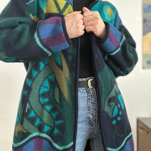 Könnte beinhalten: Eine lange, gemusterte Jacke in Marine-, Türkis-, Grün- und Lilatönen. Die Jacke hat einen Reißverschluss und ein einzigartiges geometrisches Design. Die Ärmel haben ein gestreiftes Muster in Blau und Lila. Die Jacke wird über einem schwarzen Rollkragenpullover und einer blauen Jeans getragen.