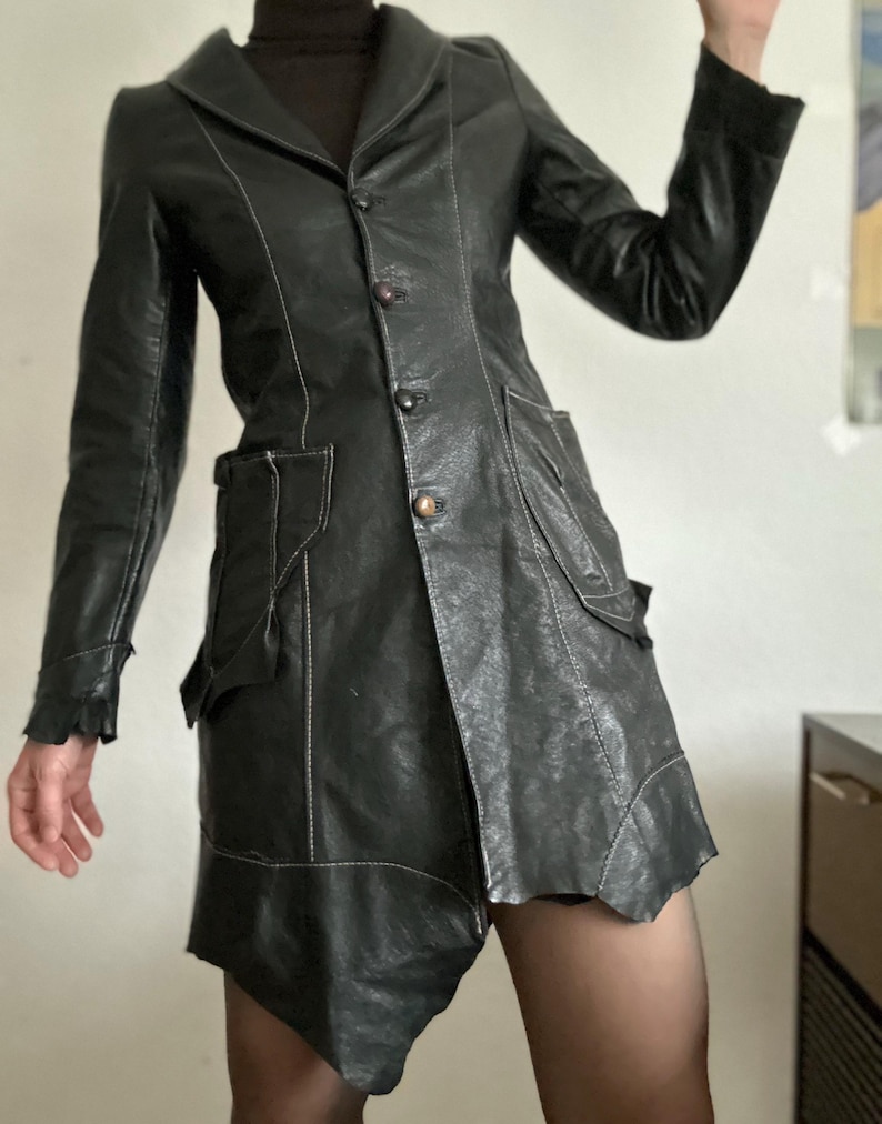 May include: Black leather jacket with a notched collar, button closure, and asymmetrical hemline. The jacket features white stitching details, two front pockets, and long sleeves. The jacket is a unique, stylish piece.