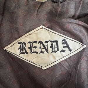 Puede incluir: Primer plano de una etiqueta en forma de rombo con el nombre "RENDA" en letras g&oacute;ticas negras. La etiqueta est&aacute; cosida sobre una tela marr&oacute;n estampada con un dise&ntilde;o floral. La tela parece ser parte del forro de una prenda.