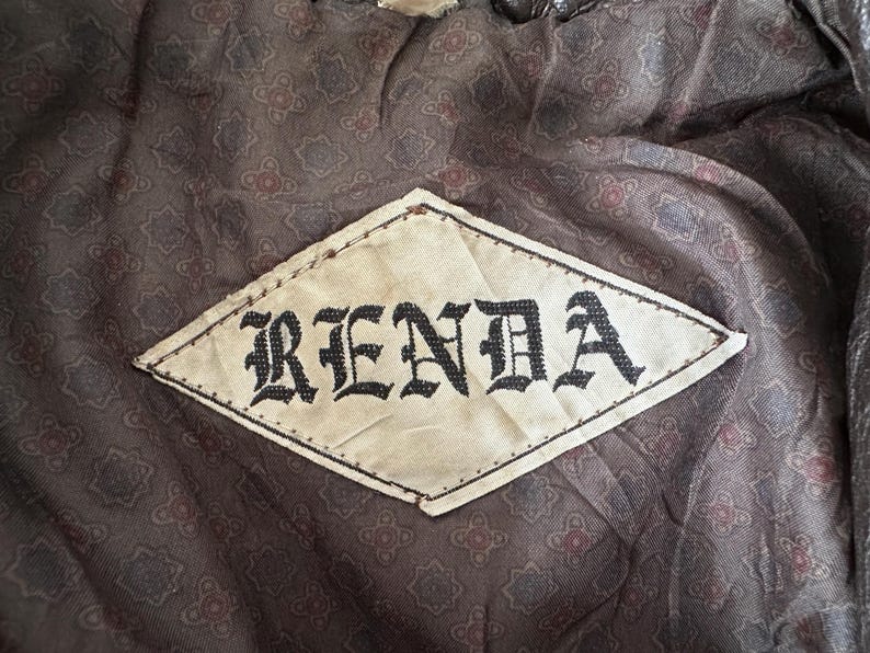 Puede incluir: Primer plano de una etiqueta en forma de rombo con el nombre "RENDA" en letras g&oacute;ticas negras. La etiqueta est&aacute; cosida sobre una tela marr&oacute;n estampada con un dise&ntilde;o floral. La tela parece ser parte del forro de una prenda.