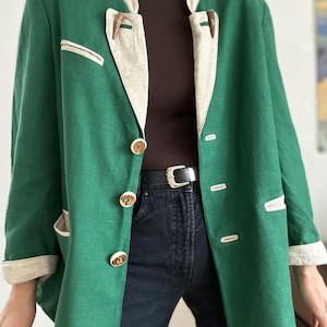 Pode incluir: Um blazer verde com acabamentos bege e fechos de alternância de madeira. O blazer tem botões dourados e um detalhe de bolso. A modelo usa um cinto preto e jeans escuros. O blazer é de estilo vintage.