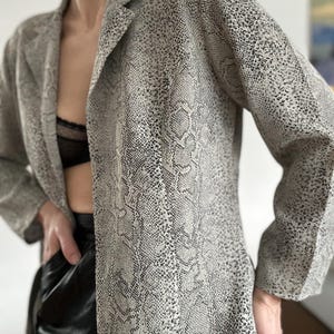 Könnte beinhalten: Ein langer Blazer mit offenem Schnitt und Schlangenmuster in Grau- und Schwarztönen. Der Blazer hat einen Reverskragen und lange Ärmel. Der Stoff scheint leicht zu sein. Der Blazer ist Teil einer Bekleidungskollektion.