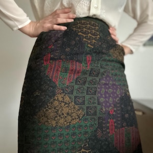 Può includere: Gonna fantasia con un design patchwork nei toni del nero, verde, viola e oro. La gonna presenta una varietà di motivi floreali e geometrici. La gonna è indossata con una camicetta bianca.