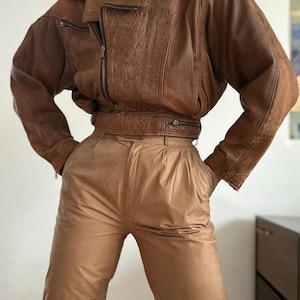 Puede incluir: Chaqueta de cuero marrón con cremallera y cuello. La chaqueta tiene un aspecto texturizado y se combina con pantalones de cuero color beige. Los pantalones tienen cintura alta y parte delantera plisada. El conjunto es de estilo vintage.
