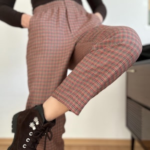 May include: Brown plaid pants with a red and black check pattern. The pants are paired with brown suede boots with decorative accents. The boots have a lug sole and lace-up closure. The outfit is completed with a long-sleeved black top.