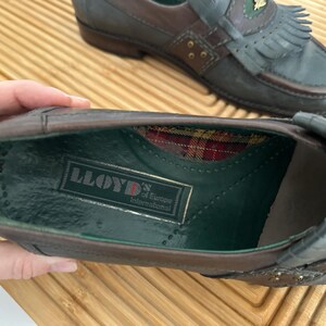 Puede incluir: Primer plano de un par de mocasines de cuero. Los zapatos son marrones y grises con flecos decorativos y detalles dorados. El interior de un zapato revela un estampado de cuadros y una etiqueta "LLOYD of Europe International".