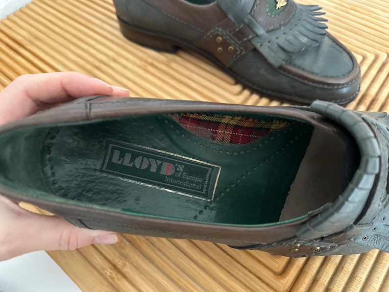 Puede incluir: Primer plano de un par de mocasines de cuero. Los zapatos son marrones y grises con flecos decorativos y detalles dorados. El interior de un zapato revela un estampado de cuadros y una etiqueta "LLOYD of Europe International".